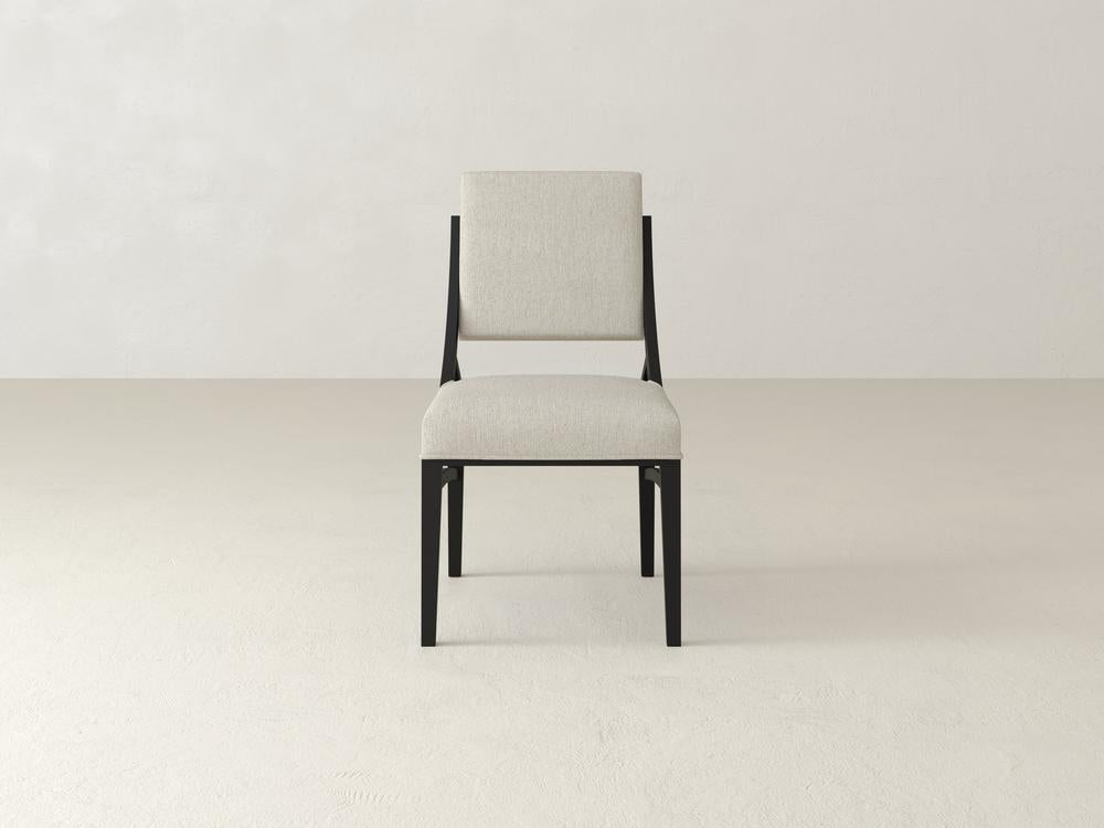 A single, modern chair with a light gray upholstered seat and backrest stands on a plain, light-colored floor against a textured, off-white wall. The chair has sleek, dark wooden legs and frame.