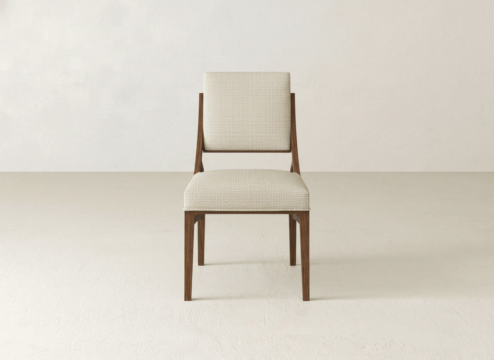 The Quinn Dining Chair by James+James features a wooden frame with light beige upholstery on the seat and backrest, ideal for elevating your dining space against simple, light-colored walls and floors.