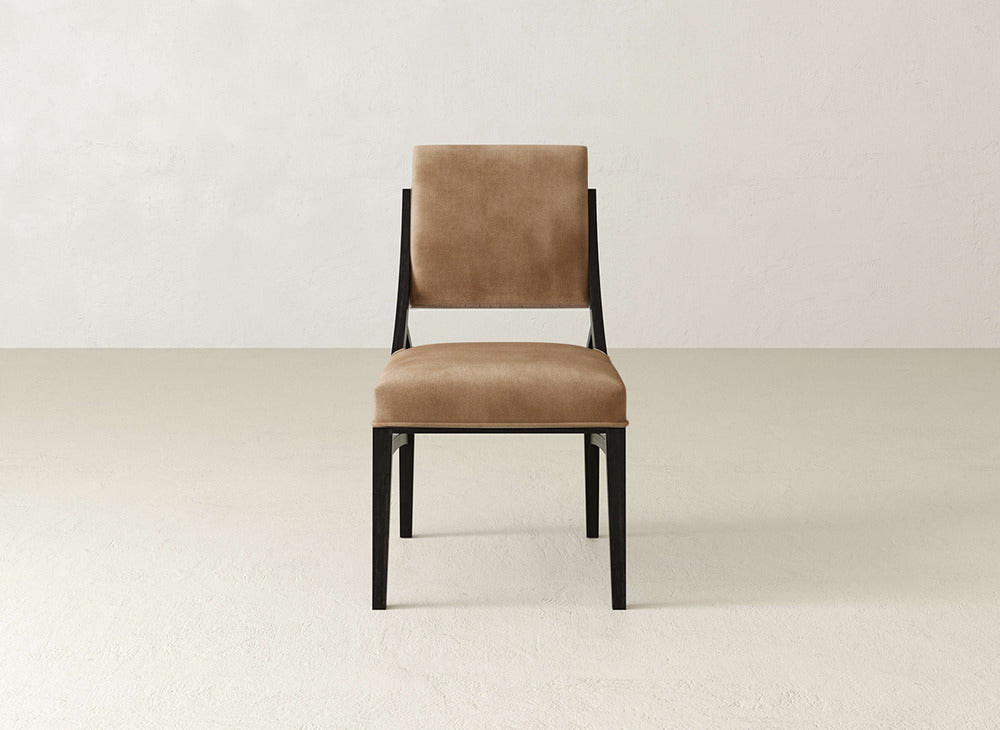 The James+James Quinn Dining Chair features a brown upholstered seat and back on a sleek black metal frame, ideal for enhancing any dining space, shown with a light beige floor and off-white wall.