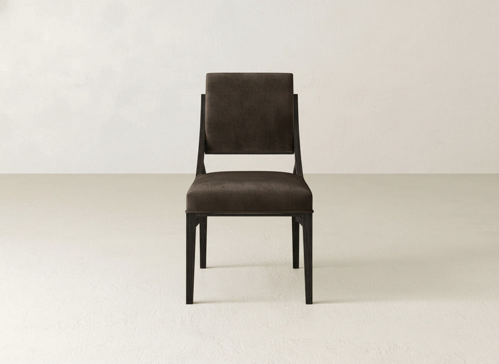 The Quinn Dining Chair by James+James features dark brown upholstery and black wooden legs, creating a sleek look that blends style and comfort in any dining room. Placed on a light floor against an off-white wall, it effortlessly enhances your décor.