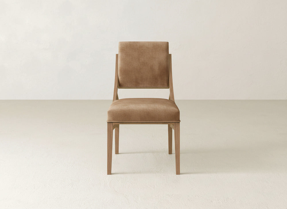 The Quinn Dining Chair by James+James, upholstered in brown with a sturdy wooden frame, is showcased in a simple, light-filled room—ideal for enhancing your dining space decor.