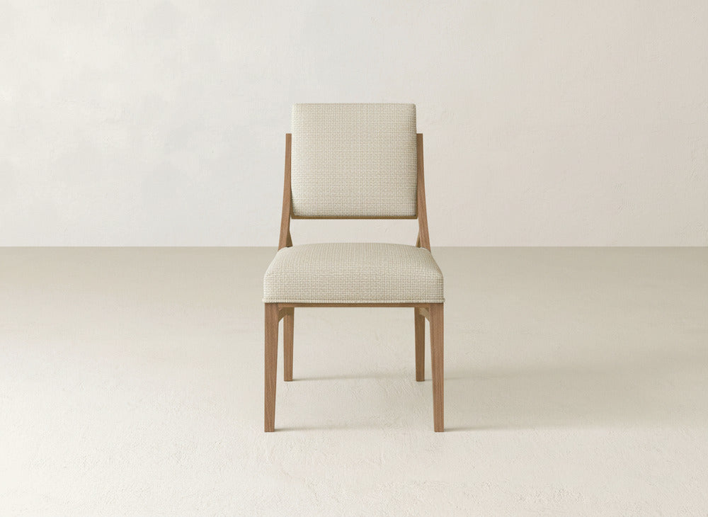 The James+James Quinn Dining Chair, featuring a light beige upholstered seat and backrest, is centered against an off-white wall and floor—an elegant minimalist addition to any modern dining room.