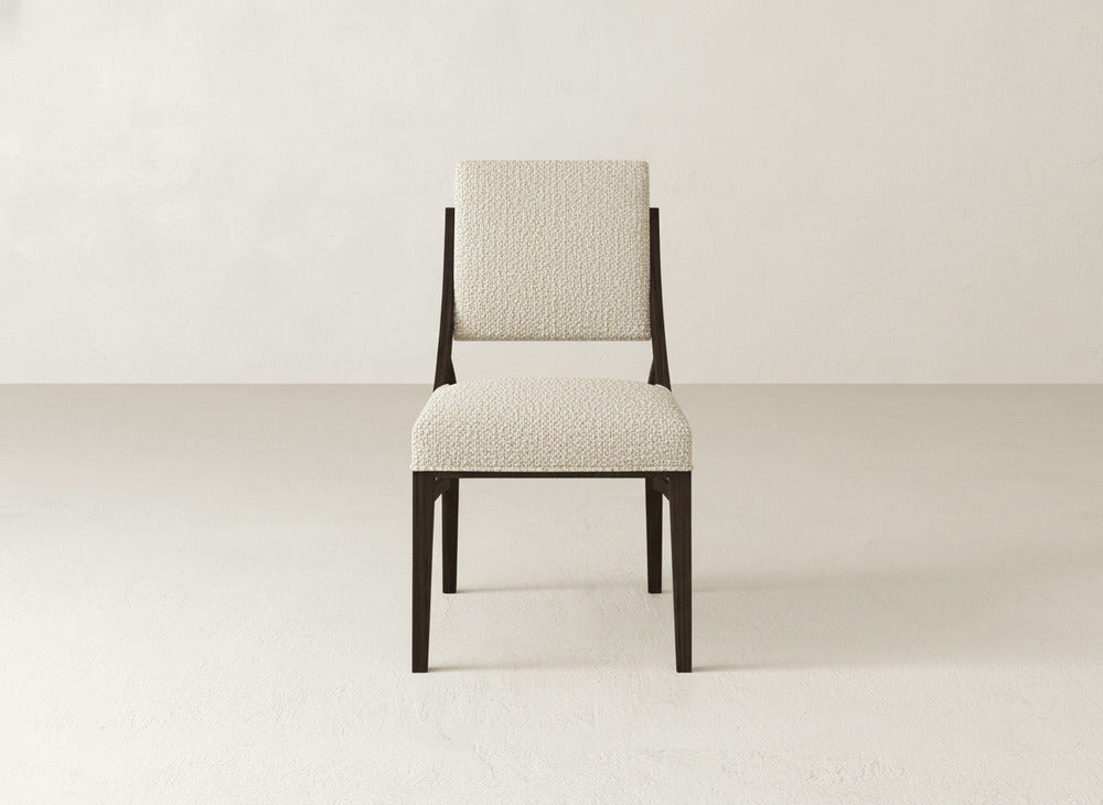 The Quinn Dining Chair by James+James features a dark wood frame and light textured upholstery, offering an elegant touch to any dining room. Displayed on a light floor against a beige wall, it enhances your decor with its refined design.