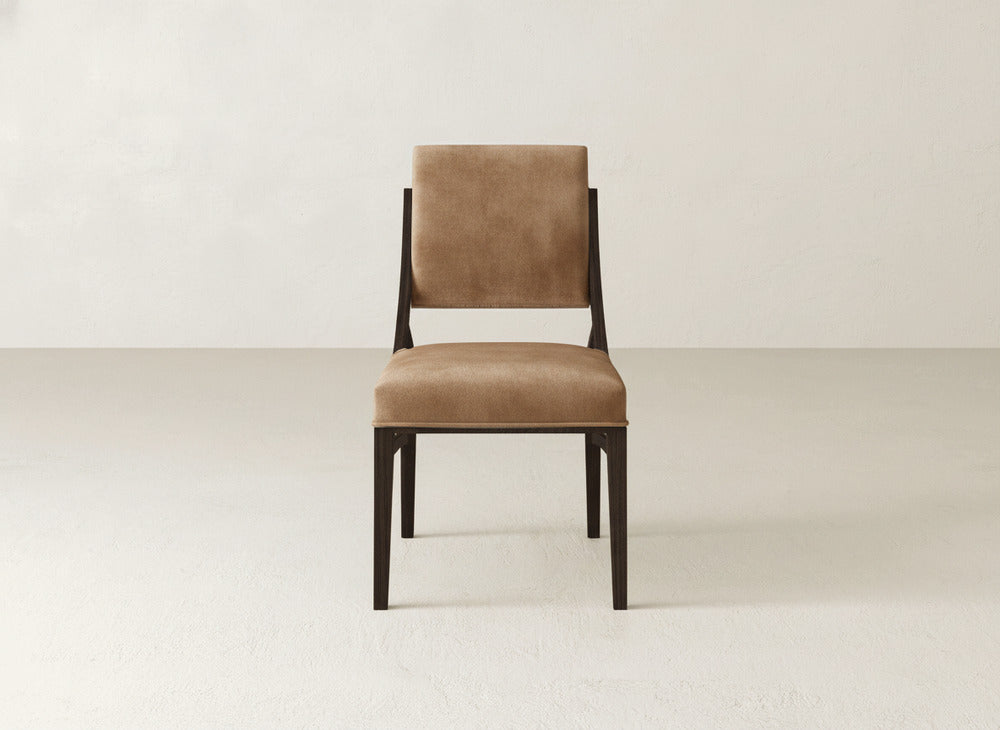 The Quinn Dining Chair by James+James features brown upholstery and a dark wood frame, shown facing forward on a light floor with an off-white wall—an ideal choice to elevate your dining room decor.