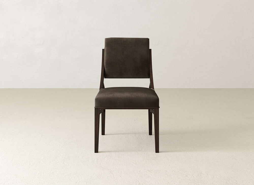 The Quinn Dining Chair by James+James features dark brown upholstery and wooden legs, with a straight backrest, set against a light wall—a sleek, modern piece to enhance any dining room.