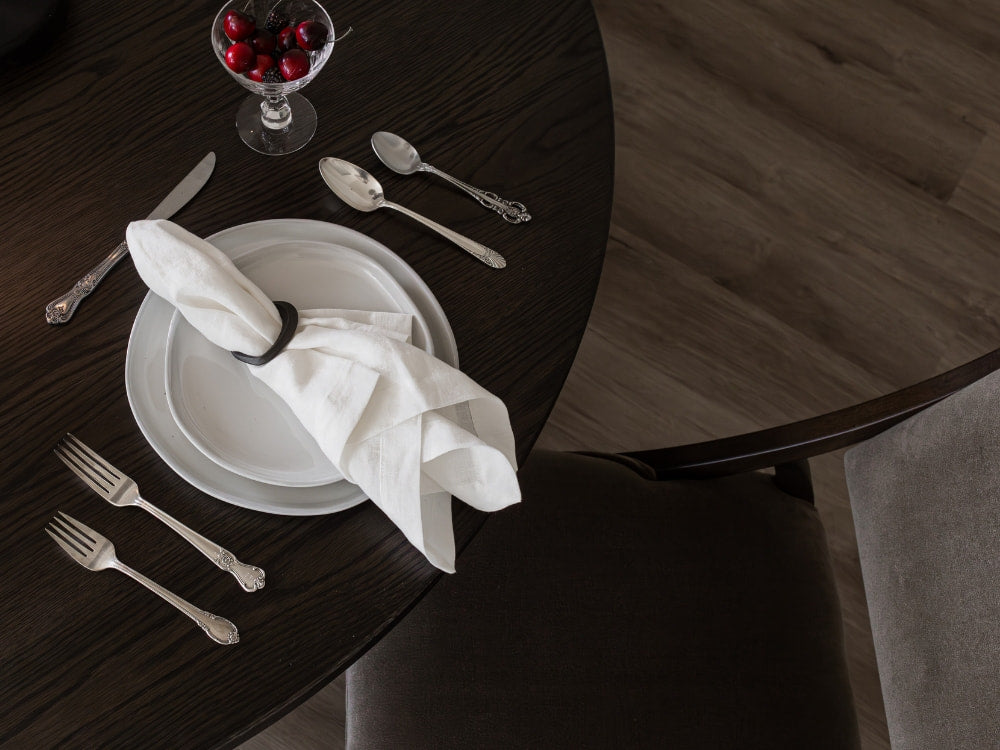A dark wood dining table set with a white plate, napkin, silverware, and glass bowl of cherries is viewed from above. Beside the table, a James+James Mink - Mohair Swatch chair adds an inviting, layered touch.