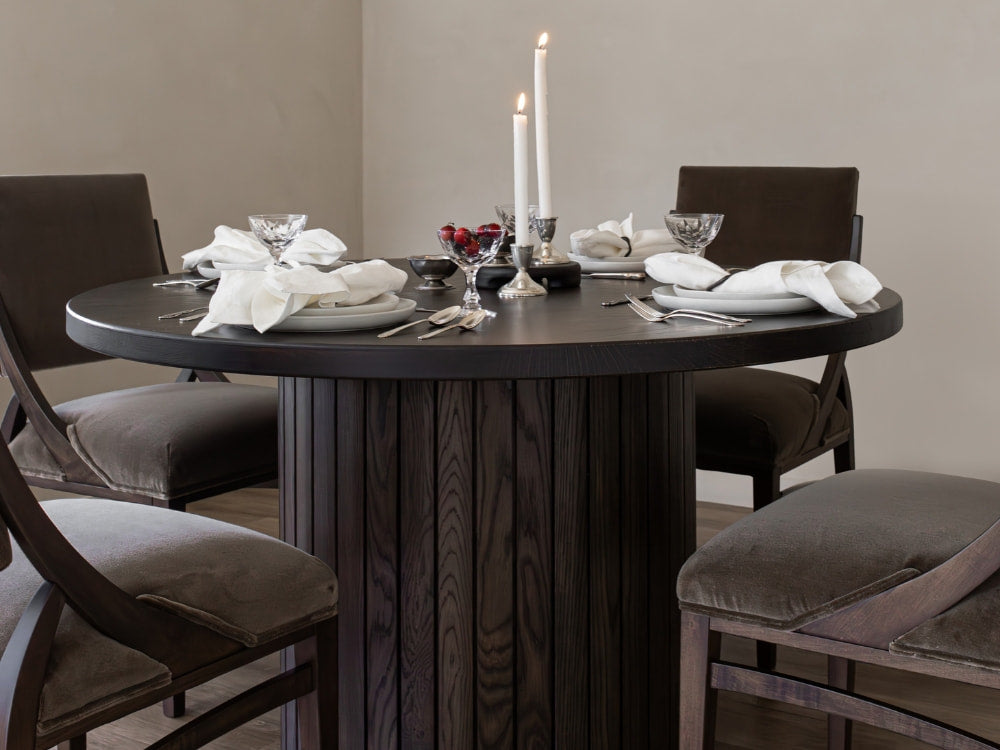 A round dark wood dining table set for four features white napkins, silverware, crystal glasses, and three lit white candles in silver holders. James+James Mink - Mohair chairs add inviting texture and a stylish layered look.