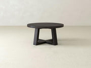 A Rae Coffee Table - Charred Ember from James+James is placed in a minimalist room with light-colored walls and floor. This round wooden table features a sturdy, geometrically designed base with intersecting legs and has a dark finish.