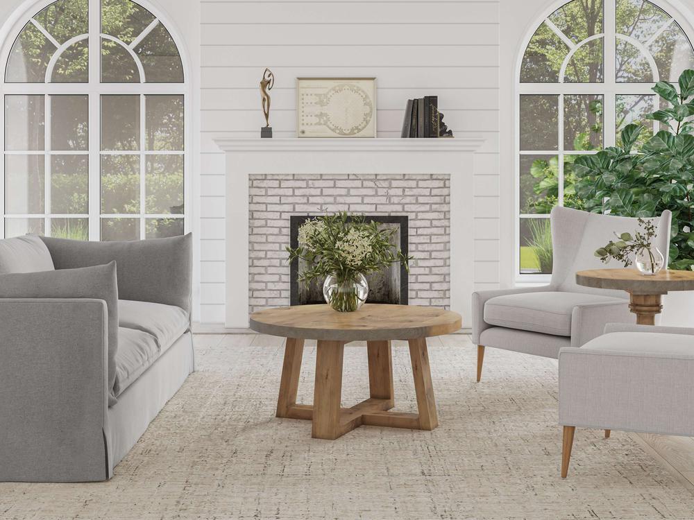 A cozy living room with a fireplace and two arched windows. The space features a gray sofa, a gray armchair, and the Rae Coffee Table by James+James in Harvest Wheat, topped with a vase of flowers. Books and decorations are arranged on the mantel and side table. Light streams in from the windows.