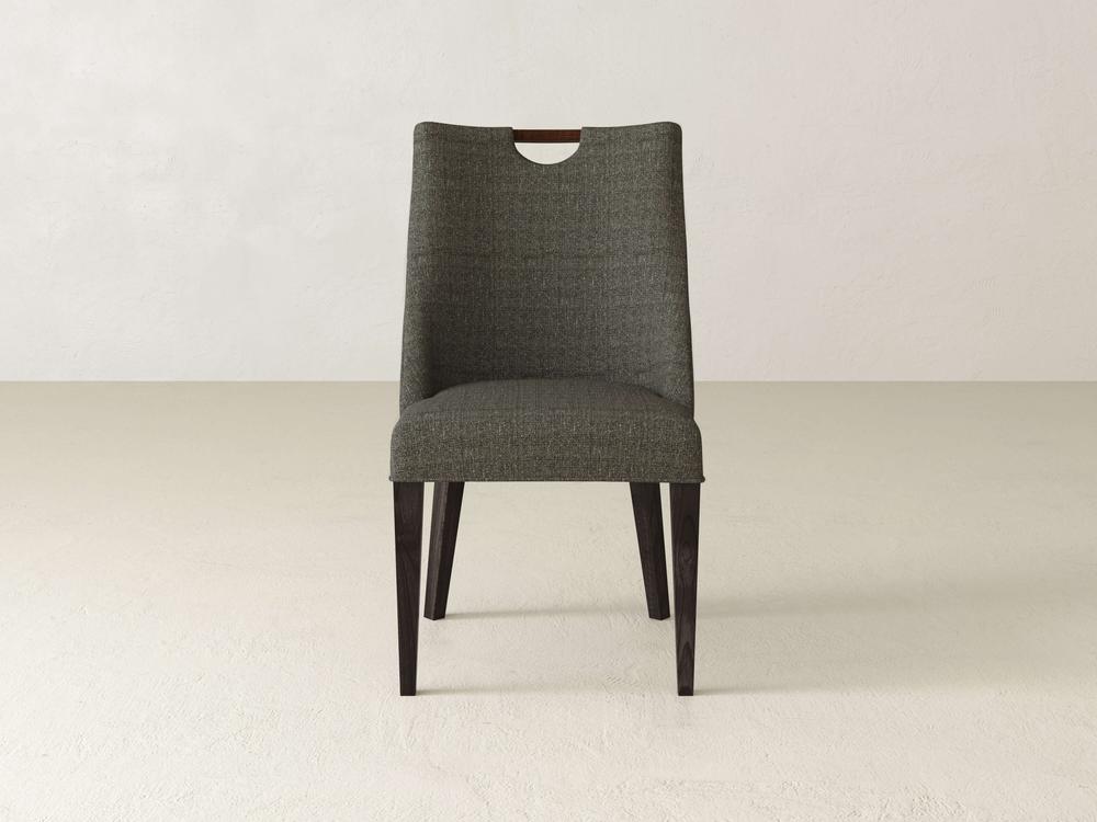 The Rosalie Dining Chair by James & James features a gray upholstered design with a sleek silhouette and wooden detail on the backrest, standing elegantly on a light-colored floor against a white wall, complemented by modern dark wooden legs.