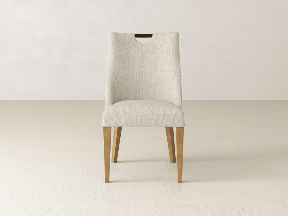 The Rosalie Dining Chair by James & James features light beige upholstery and wooden legs, showcased on a light floor against a neutral wall. Its modern elegance is highlighted by a slightly curved backrest and a top cutout.