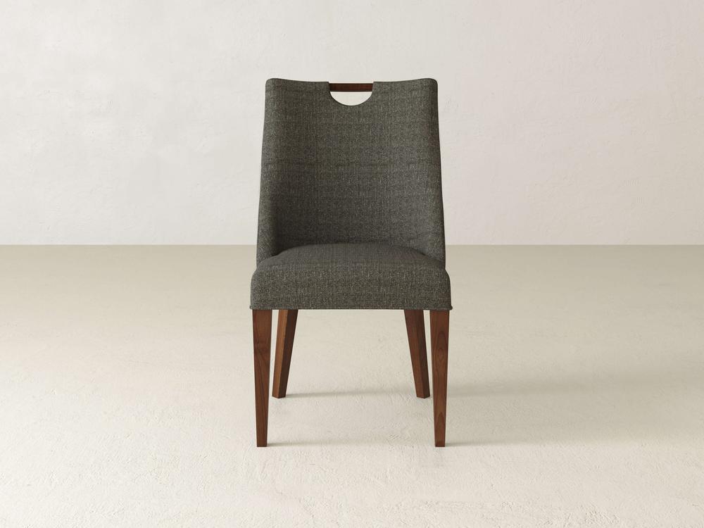 The Rosalie Dining Chair by James & James showcases modern elegance with its dark gray upholstery and wooden legs. It features a slightly curved backrest and a unique wooden handle cut-out at the top, ideal for placement against a plain white wall.