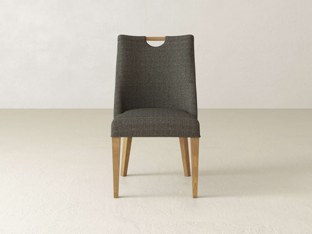 The James & James Rosalie Dining Chair exudes modern elegance with dark gray upholstery and light wooden legs, perfect for any minimalist dining setting. It features a small cut-out handle at the backrest top, adding a sophisticated touch.