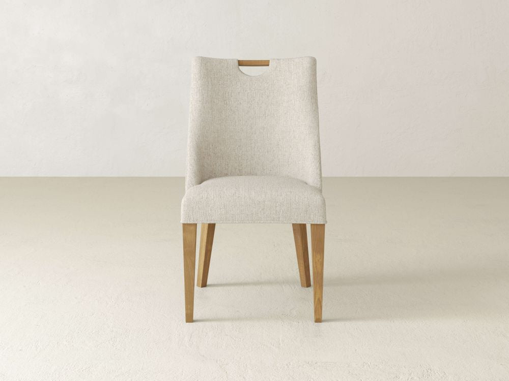 The Rosalie Dining Chair by James & James exudes modern elegance with light gray upholstery, wooden legs, and a subtle backrest cutout. It seamlessly blends into any decor, poised on a smooth, light-colored floor against a plain wall.