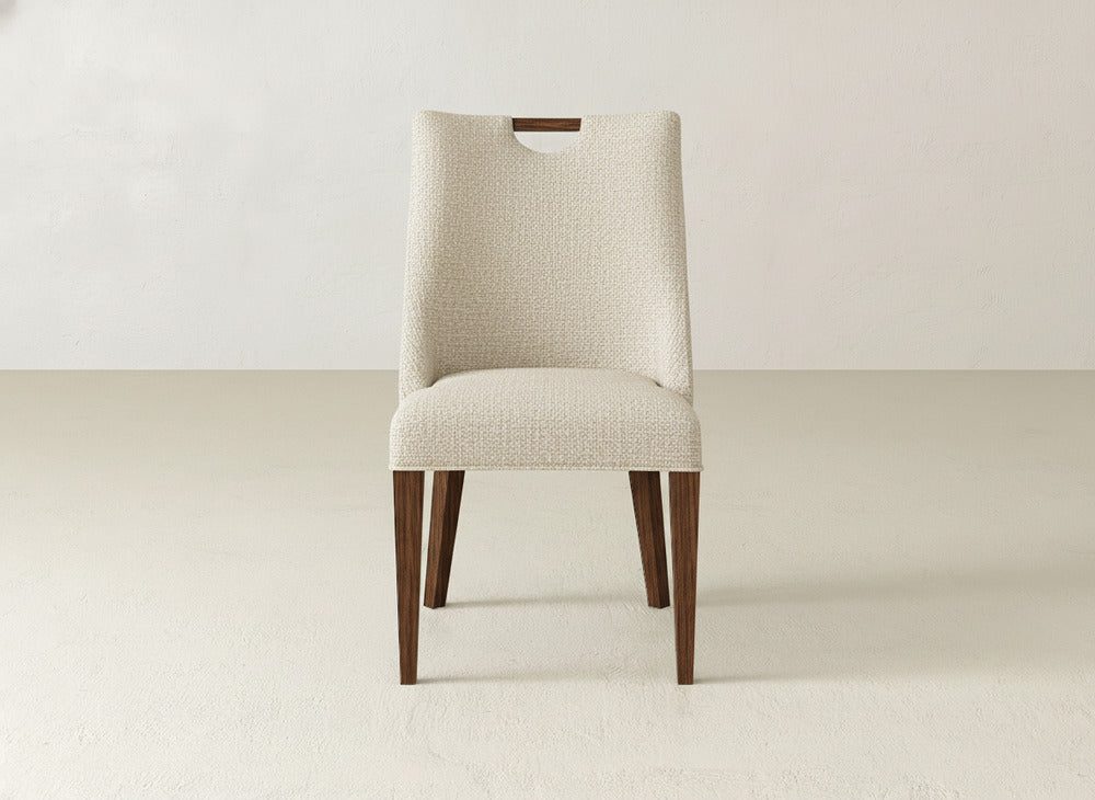 The James & James Rosalie Dining Chair features modern beige upholstery, dark wooden legs, and a small rectangular cutout at the top of the backrest, perfect for minimalist rooms with light walls and flooring.