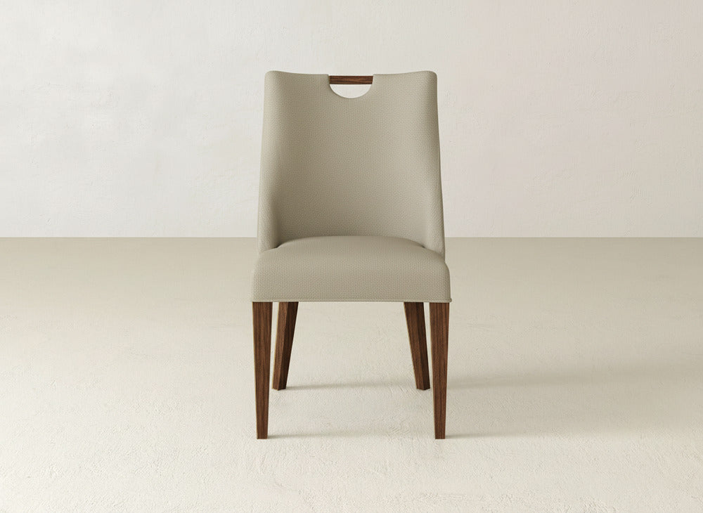 The James & James Rosalie Dining Chair features beige upholstery, dark wood legs, and a curved cutout at the top—an elegant choice for minimalist spaces with light walls and floors.