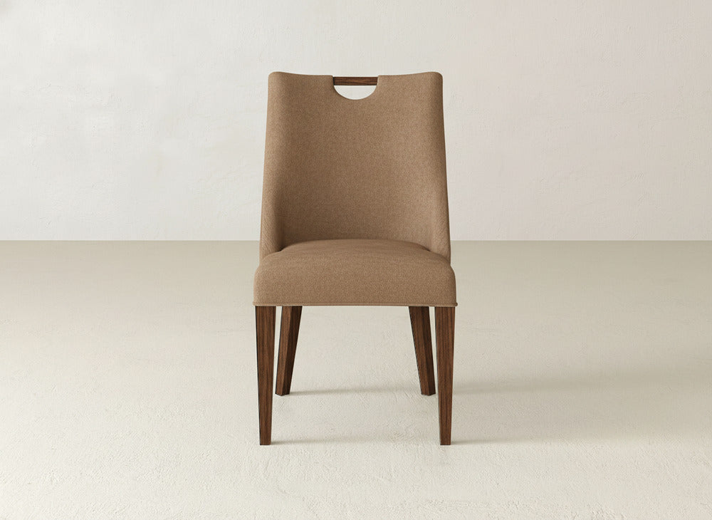 The Rosalie Dining Chair by James & James features brown upholstery, wooden legs, a high backrest, and a small top cutout—an elegant addition to minimalist rooms with light floors and plain white walls.