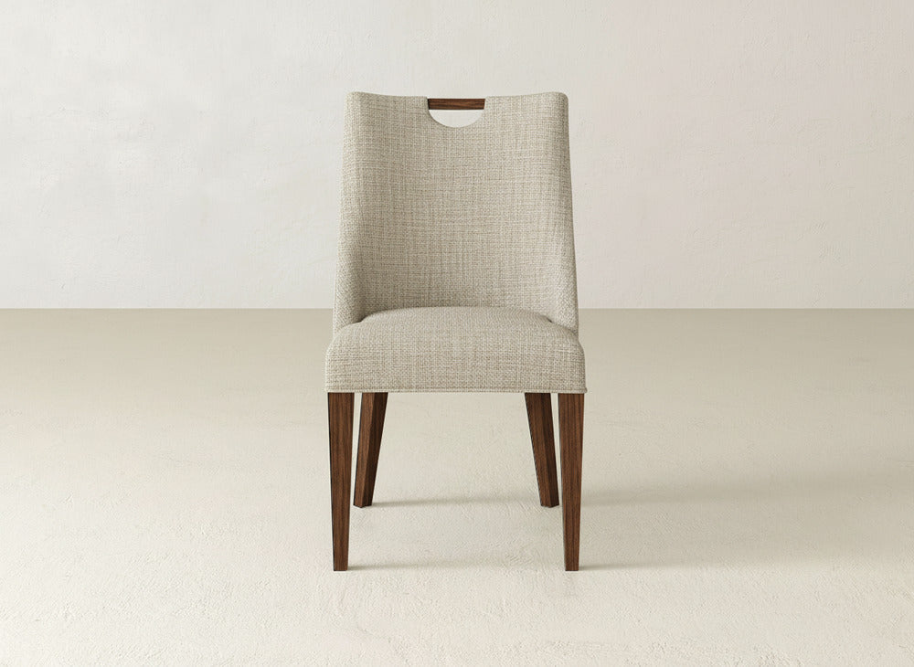 The James & James Rosalie Dining Chair features light beige fabric, a rectangular backrest cutout, and four wooden legs, offering an elegant seating option against a simple, light background.