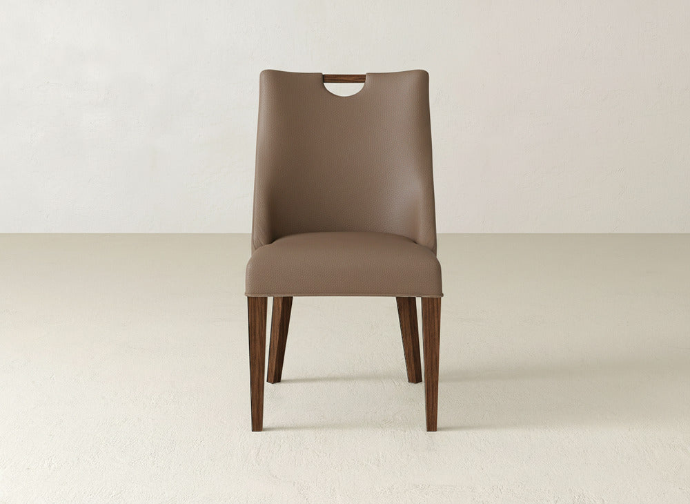 The Rosalie Dining Chair by James & James is a modern brown chair with a curved backrest, cutout handle, and wooden legs, shown on a light floor against a plain light wall.