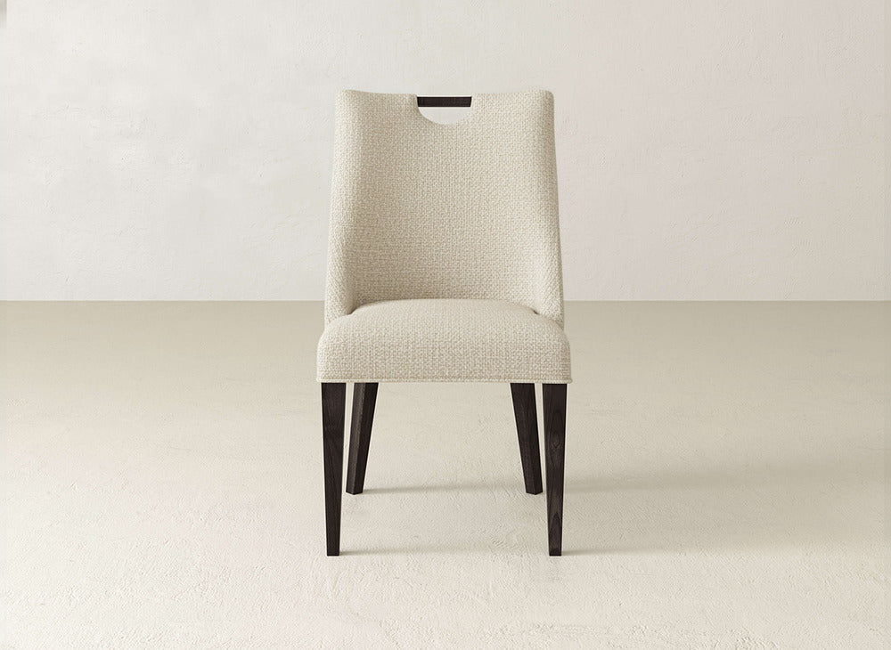 The James & James Rosalie Dining Chair features light beige upholstery, dark wood legs, and a small rectangular cut-out in the backrest—a perfect modern accent against an off-white background.