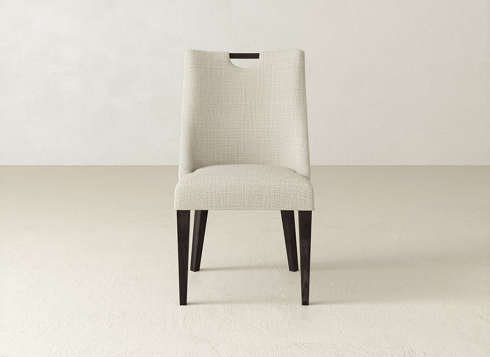 The James & James Rosalie Dining Chair pairs light beige textured upholstery with dark wooden legs and a rectangular cutout handle, offering modern style and practicality that complements any neutral space.