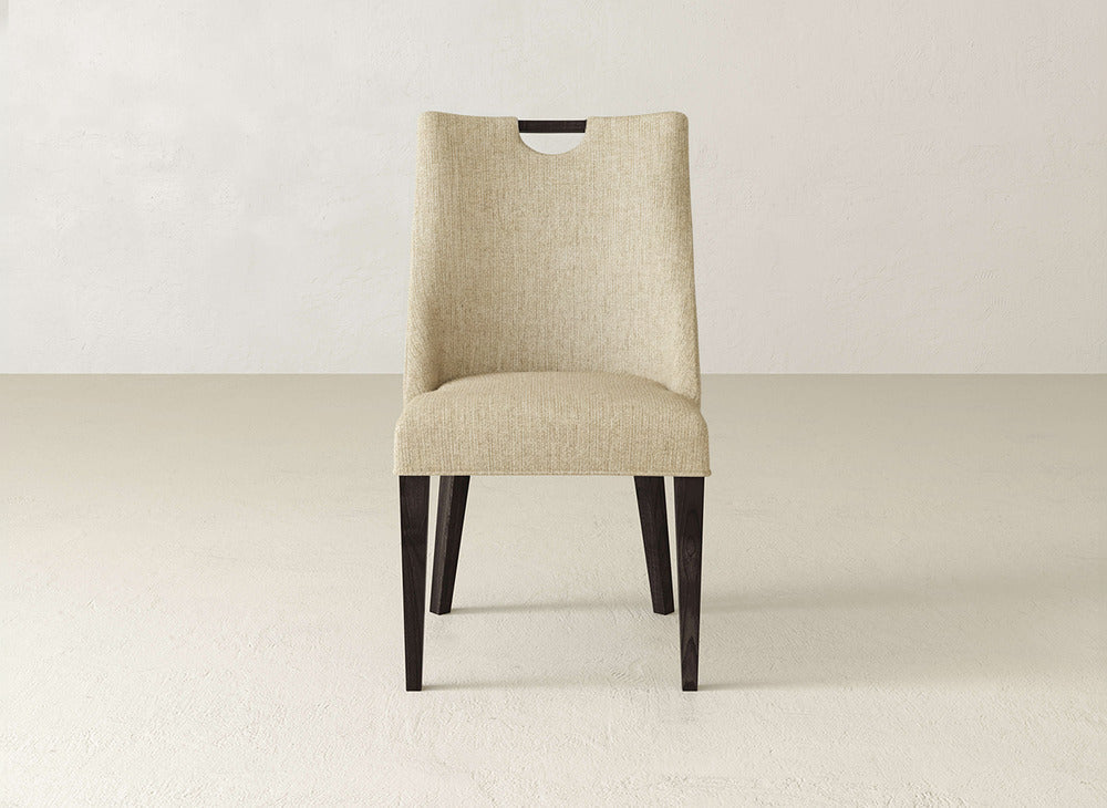 The James & James Rosalie Dining Chair features beige upholstery, dark wooden legs, and a top cutout handle on the backrest for easy movement, making it a modern addition to any dining space.