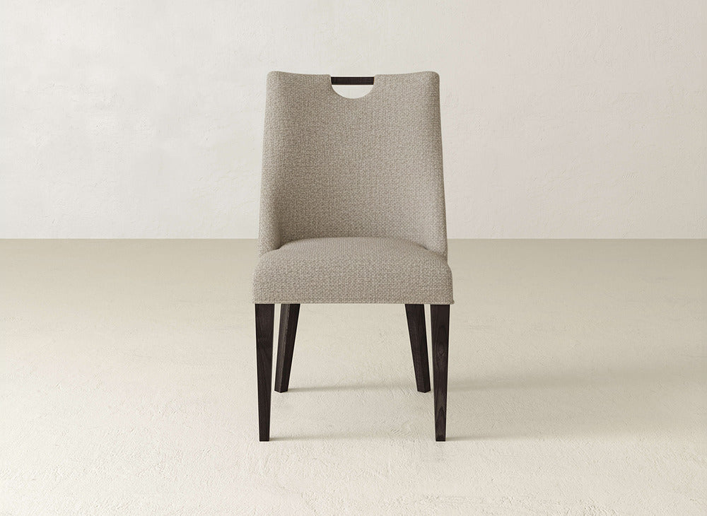 The James & James Rosalie Dining Chair features light beige upholstery, dark wood legs, and a curved backrest cutout, blending modern style with minimalist charm—perfect for neutral-toned spaces.