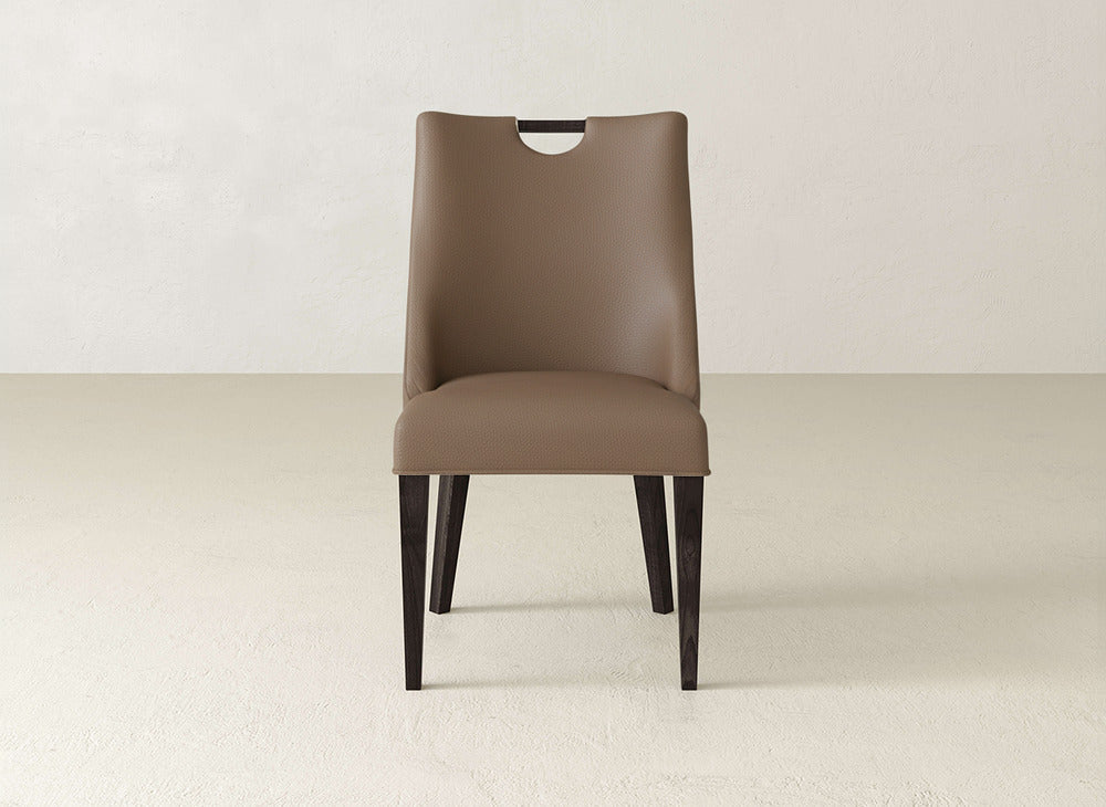 The Rosalie Dining Chair by James & James features modern brown leather, dark wooden legs, and a rounded cutout at the top, shown on a light floor and an off-white wall.