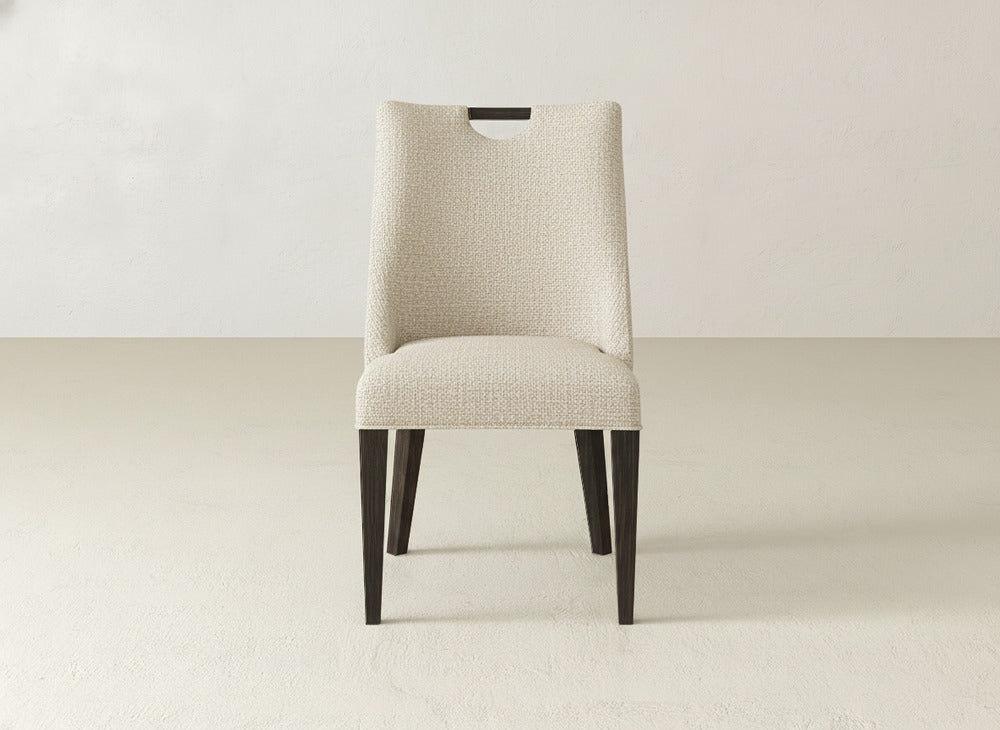 The James & James Rosalie Dining Chair features light beige upholstery, dark wood legs, and a wooden handle cut-out on the backrest, shown against a plain, light background.