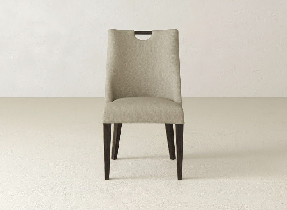 The Rosalie Dining Chair by James & James is a modern beige chair with dark wooden legs, a curved back, and a cut-out handle, shown in a minimal, light-toned room.