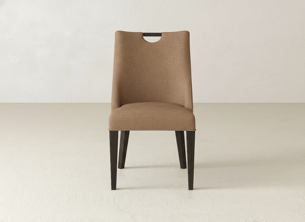 The Rosalie Dining Chair by James & James showcases brown upholstery, a straight backrest with cut-out handle, and dark wooden legs, styled on a light beige floor against a plain wall.