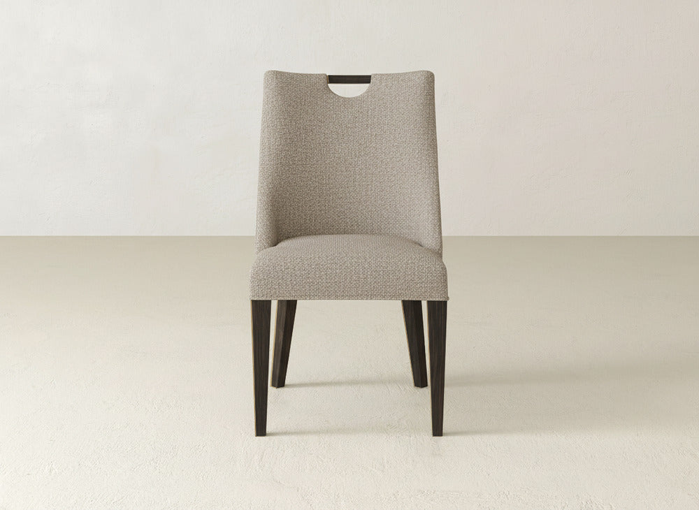 The Rosalie Dining Chair by James & James features light beige upholstery, dark wooden legs, and a sleek cutout wooden handle at the top of the backrest.