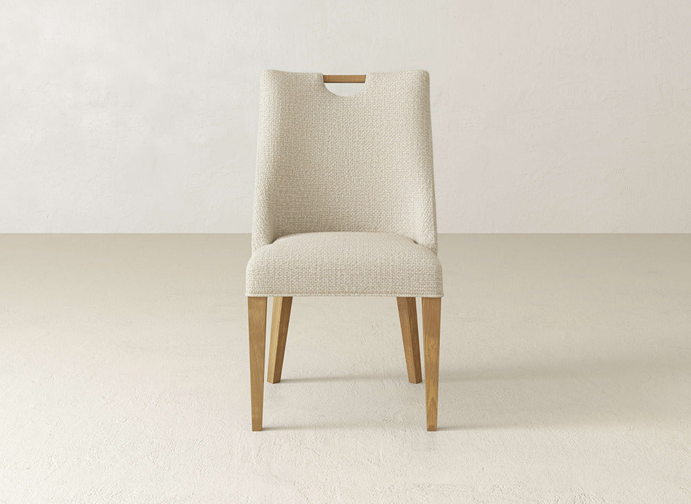 The James & James Rosalie Dining Chair features light beige textured upholstery, wooden legs, and a top cutout handle, offering an elegant look for any dining space.