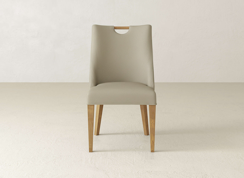 The James & James Rosalie Dining Chair features modern light beige upholstery, a curved backrest with a cut-out handle, and wooden legs, shown against a plain light-colored background.