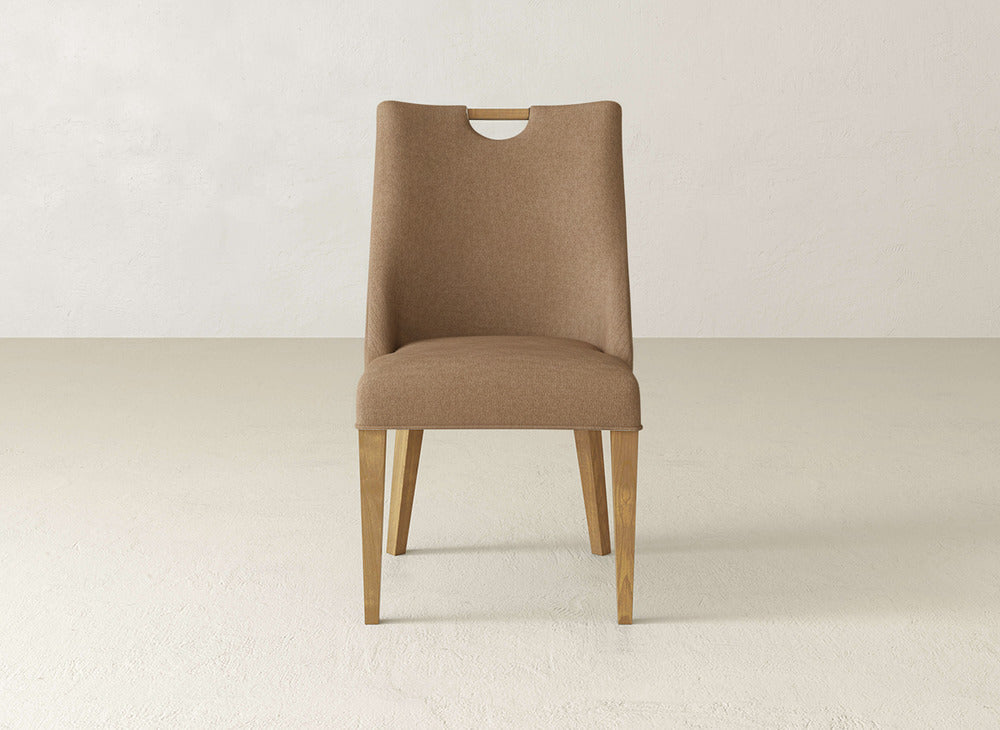 The James & James Rosalie Dining Chair features brown upholstery, light wooden legs, and a rounded top cutout, making it a modern choice for minimal, neutral-toned spaces.