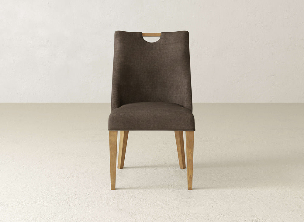 The Rosalie Dining Chair by James & James features light wooden legs, a dark brown upholstered seat and back with a cutout handle near the top, all set against a plain light-colored background.