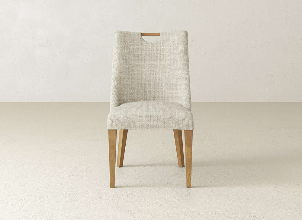 The James & James Rosalie Dining Chair features light beige upholstery, wooden legs, and a small rectangular cutout atop the backrest, creating an elegant look against a plain, light background.