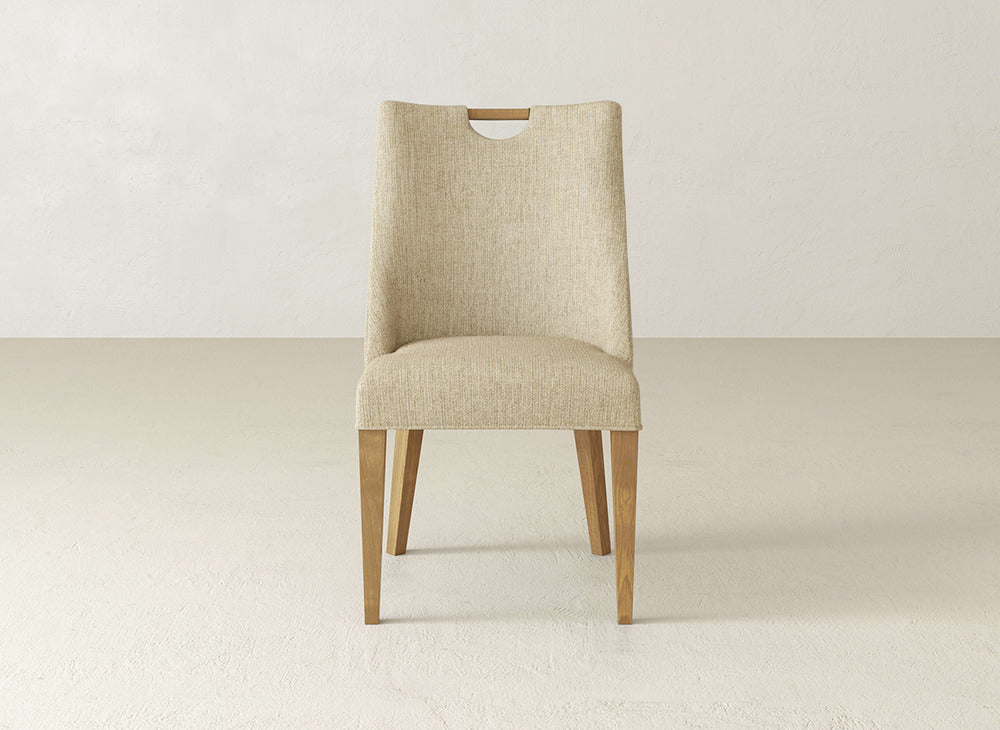 The James & James Rosalie Dining Chair features beige upholstery, light wooden legs, and a small handle cutout on the backrest, shown against a plain light wall and floor.