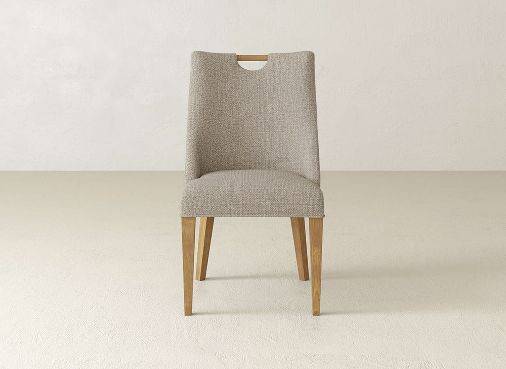 The Rosalie Dining Chair by James & James features a modern design with a light beige upholstered seat and backrest, curved cutout at the top, and wooden legs, set against a neutral background.