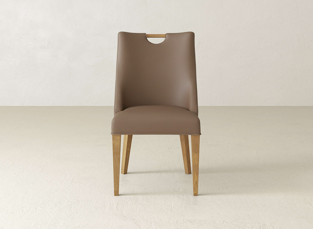 The Rosalie Dining Chair by James & James features brown upholstery, wooden legs, a straight back with a small oval cutout at the top, and sits against a plain white wall on a light-colored floor.