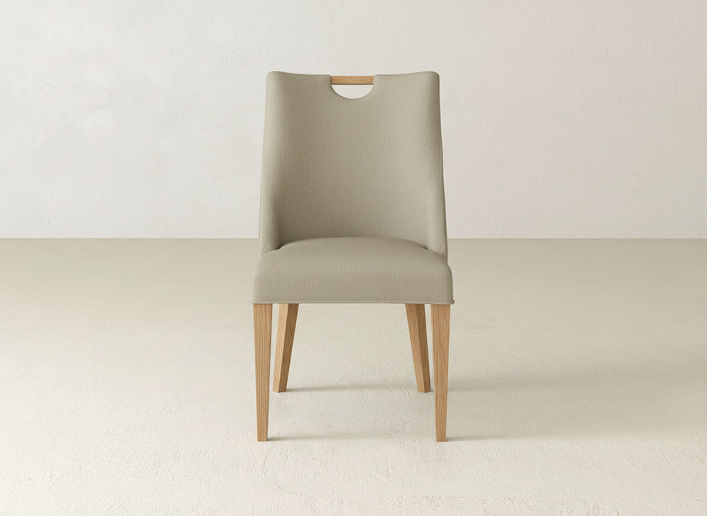 The James & James Rosalie Dining Chair, upholstered in beige with wooden legs, stands at the center of a minimalist room with light-colored walls and flooring.