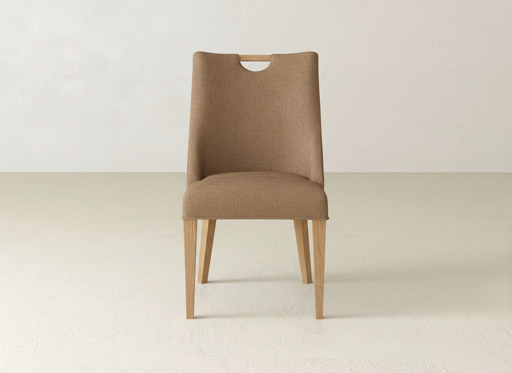 The James & James Rosalie Dining Chair has light wooden legs, a light brown upholstered seat with a cut-out handle, and offers an elegant seating option for modern spaces. It’s shown against a plain, light wall and floor.