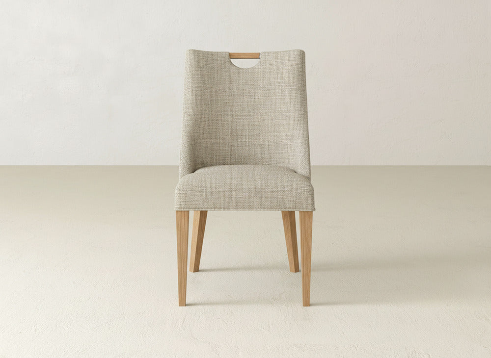 The Rosalie Dining Chair by James & James features light wooden legs and a light gray upholstered seat and backrest with a small rectangular cutout at the top, shown in a minimalist room with light floors and walls.