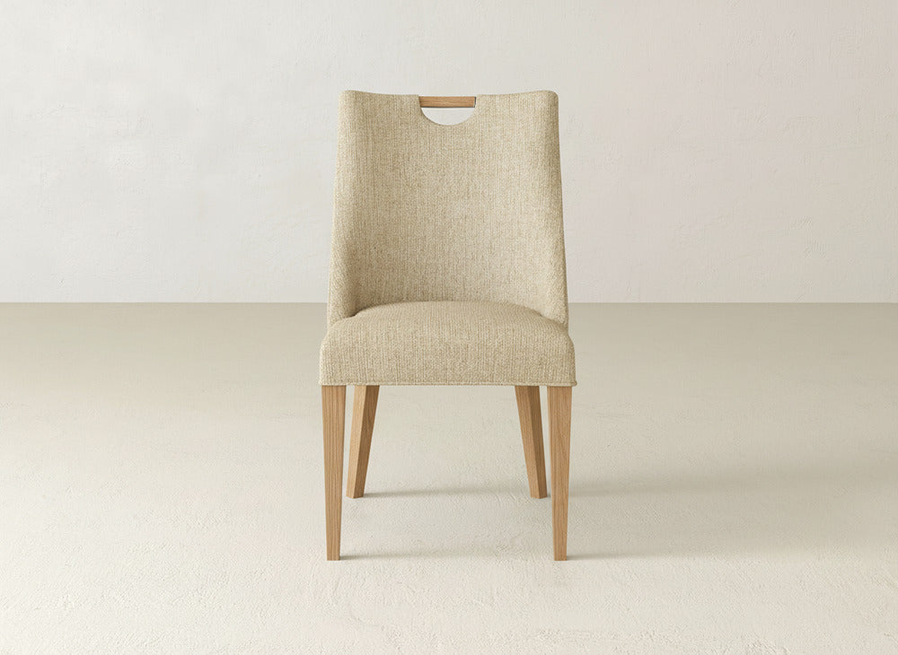 A James & James Rosalie Dining Chair, upholstered in beige with light wood legs, rests on a pale floor against an off-white wall, facing forward.