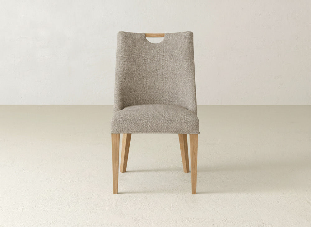 The Rosalie Dining Chair by James & James is a modern piece with light wooden legs, textured beige upholstery, and a small rounded cutout at the top of the seat back, shown against a light background.