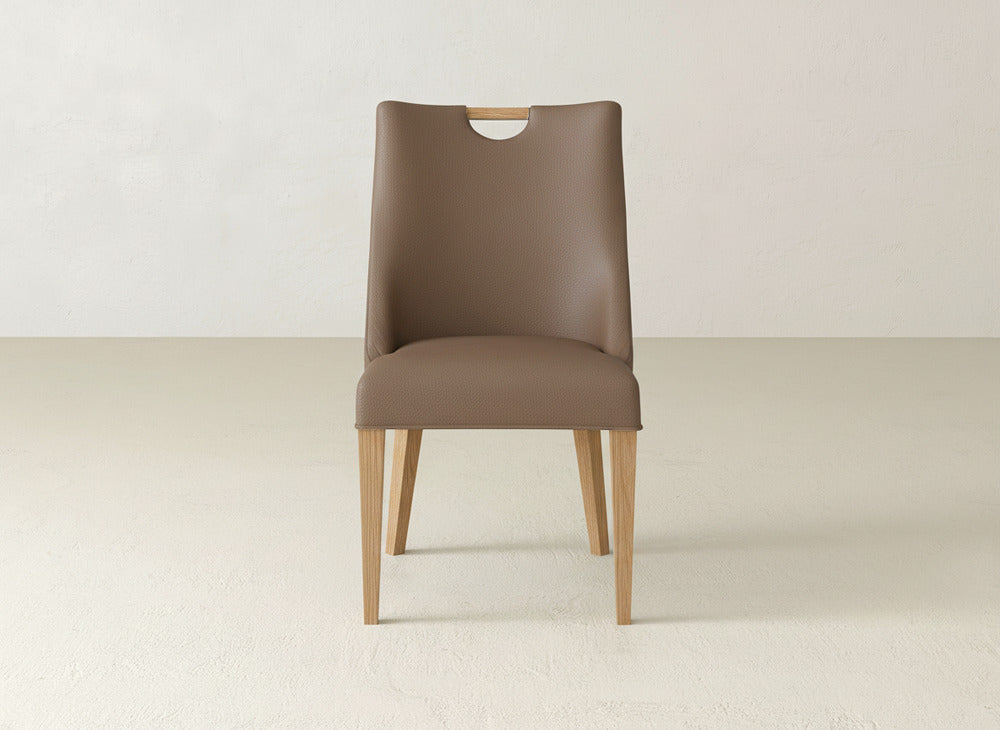 The James & James Rosalie Dining Chair, featuring light wooden legs and a cutout handle atop the backrest, sits in a neutral room, seamlessly blending modern design with understated elegance.