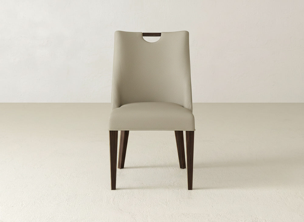 The James & James Rosalie Dining Chair is a modern piece with a curved backrest and top cut-out, dark wooden legs, and looks elegant on a light textured floor near a plain light-colored wall.