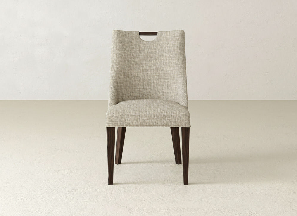 A modern beige James & James Rosalie Dining Chair with dark wood legs faces forward in an empty, light-colored room with a plain wall and floor.