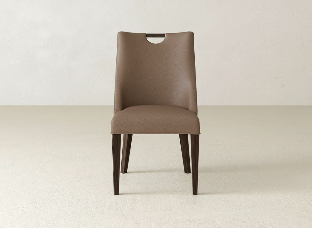 The James & James Rosalie Dining Chair features brown upholstery, dark wooden legs, and a rounded cutout near the top of the backrest, shown against an off-white wall.