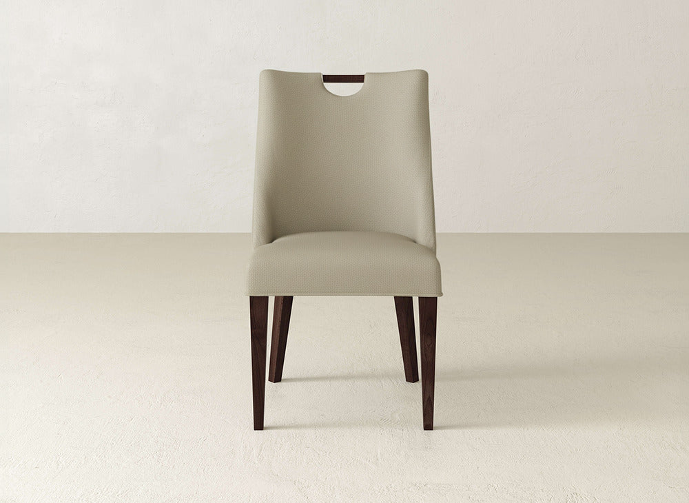 The Rosalie Dining Chair by James & James features beige upholstery, dark wood legs, and a curved backrest with a wooden handle cutout, perfect for minimalist, light-colored spaces.